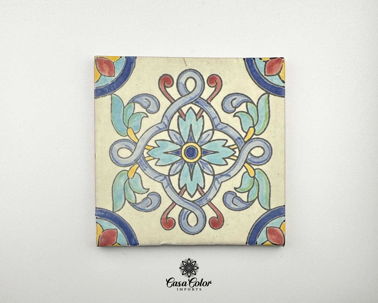 handmade tile 6x6 inches. The tile is painted by hand with a brush. The colors are a white background with a blue flower in the center. The flower has long branches coming out from the flower going in different directions. The color of the branches are lighter blue, red and yellow. The flower in the center has yellow, with light blue petals. 